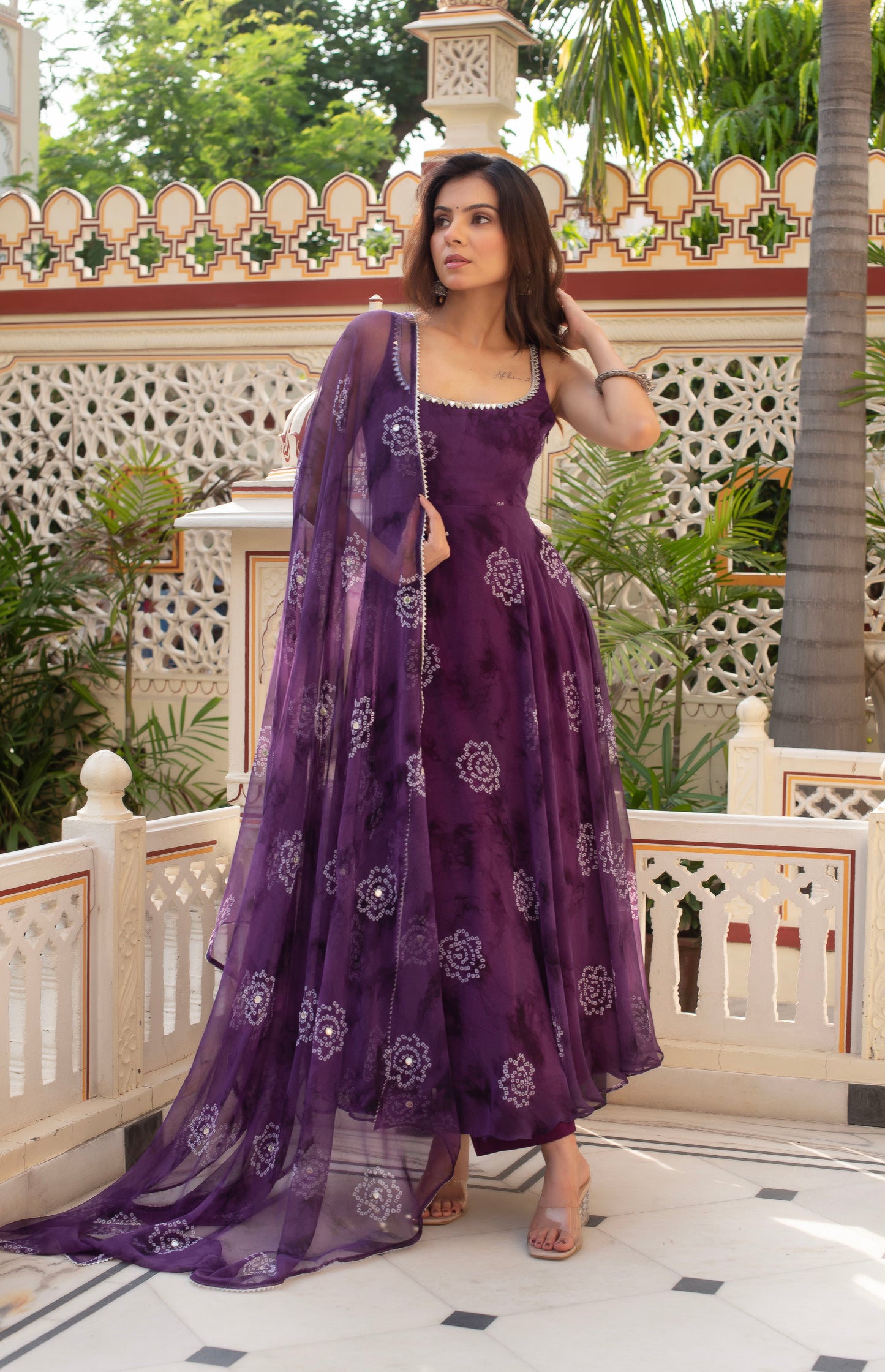 Woman in a purple traditional outfit standing in an outdoor setting with decorative walls and plants.